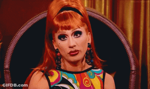 A Drag Queen Is Sitting In A Chair Wearing A Colorful Shirt And Earrings . GIF