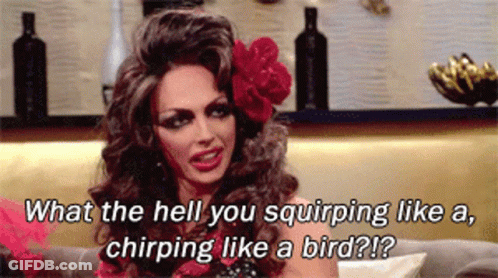 A Drag Queen Is Sitting On A Couch With A Flower In Her Hair GIF