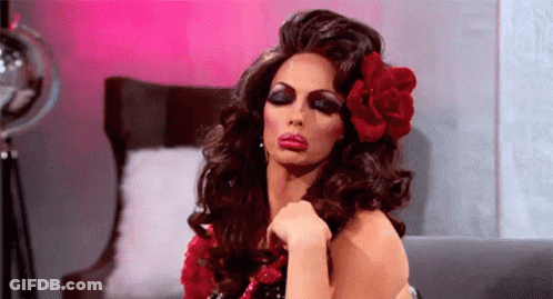 A Drag Queen Is Sitting On A Couch With A Red Flower In Her Hair . GIF
