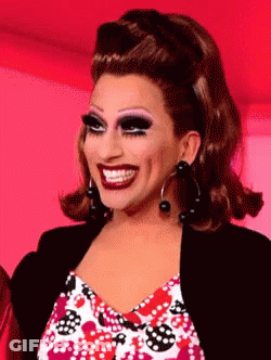 A Drag Queen Is Smiling And Wearing A Black Jacket And Earrings GIF
