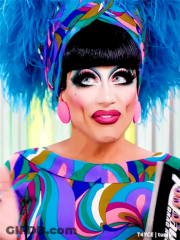 A Drag Queen Is Wearing A Colorful Dress And Feathered Headpiece GIF