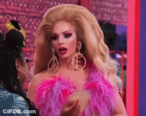 A Drag Queen Is Wearing A Pink Feathered Top And Earrings And Talking To Another Drag Queen . GIF