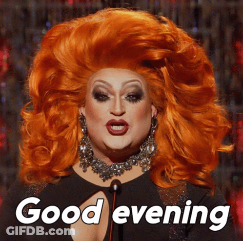 A Drag Queen Says " Good Evening " While Wearing A Large Red Wig GIF