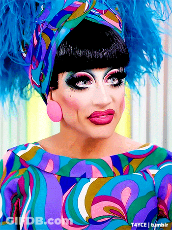 A Drag Queen Wearing A Colorful Dress And Feathered Headpiece GIF