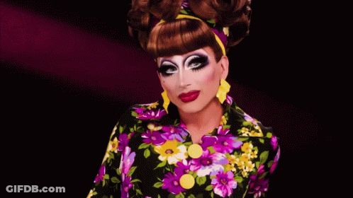 A Drag Queen Wearing A Colorful Floral Shirt And Yellow Earrings GIF