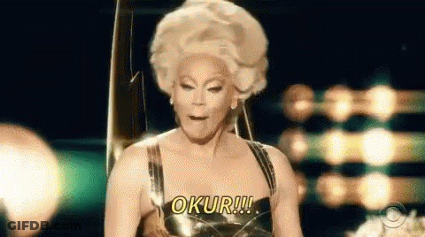 A Drag Queen Wearing A Wig And A Gold Dress Is Making A Funny Face While Standing On A Stage . GIF