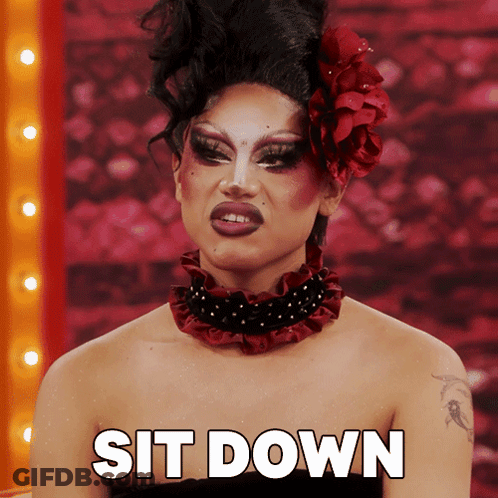 A Drag Queen With A Red Flower In Her Hair Is Sitting Down GIF