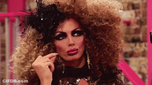 A Drag Queen With A Very Large Curly Hair And A Black Hat GIF