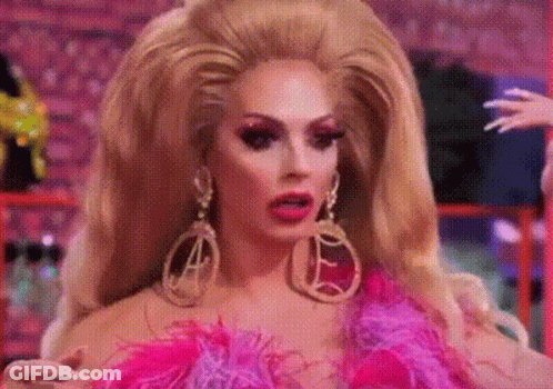 A Drag Queen With A Very Large Wig And Earrings Is Wearing A Pink And Purple Outfit . GIF