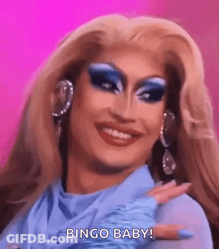 A Drag Queen With Blonde Hair And Blue Makeup Is Smiling And Making A Bingo Baby Gesture . GIF
