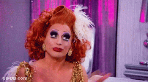 A Drag Queen With Red Hair And Feathers On Her Head Is Wearing A Gold Dress And Earrings . GIF