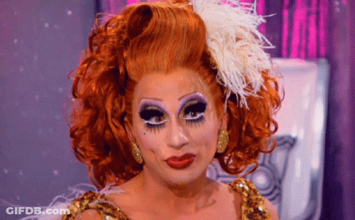 A Drag Queen With Red Hair And Feathers On Her Head Looks At The Camera GIF