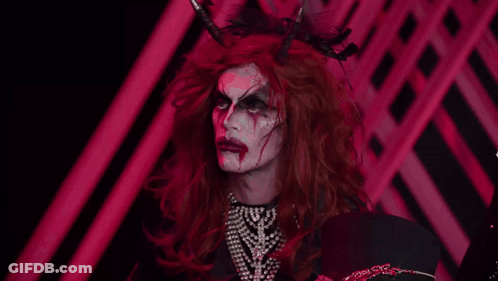 A Drag Queen With Red Hair And Horns Is Wearing A Pearl Necklace GIF