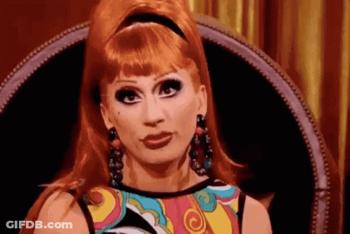 A Drag Queen With Red Hair Is Sitting In A Chair And Making A Funny Face . GIF