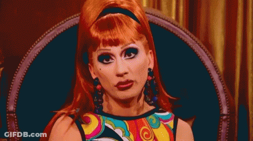 A Drag Queen With Red Hair Is Sitting In A Chair Wearing A Colorful Dress And Earrings . GIF