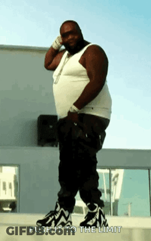 A Fat Man In A White Tank Top And Black Pants Is Standing On A Sidewalk With The Words Push It To The Limit Above Him GIF