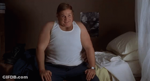 A Fat Man Is Sitting On A Bed Wearing A White Tank Top And A Watch . GIF