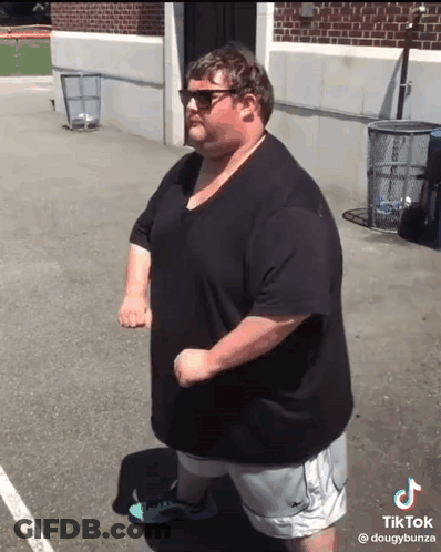 A Fat Man Wearing Sunglasses And A Black Shirt Is Standing In Front Of A Brick Building .. GIF