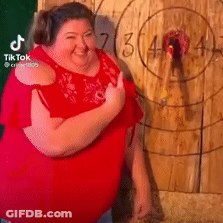 A Fat Woman In A Red Shirt Is Standing In Front Of A Wooden Target . GIF