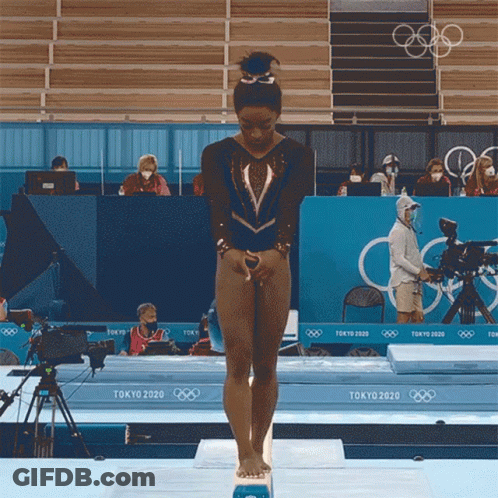 A Female Gymnast Stands On A Balance Beam In Front Of A Tokyo 2020 Sign GIF