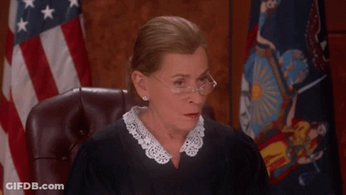 A Female Judge Wearing Glasses And A Lace Collar Sits In Front Of An American Flag GIF
