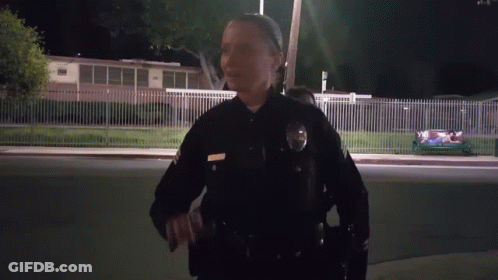 A Female Police Officer Stands In Front Of A Fence And A Building At Night GIF