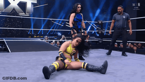 A Female Wrestler Is Laying On The Ground In A Wrestling Ring While Another Wrestler Stands Behind Her GIF
