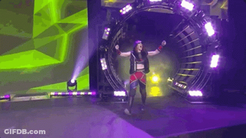 A Female Wrestler Is Walking Through A Tunnel On A Stage With Her Arms In The Air . GIF