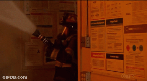 A Fireman Is Spraying Smoke In Front Of A Wall With Posters On It Including One That Says Contact Us GIF