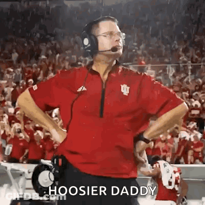 A Football Coach Wearing Headphones And A Red Shirt Is Standing On The Sidelines Of A Field . GIF