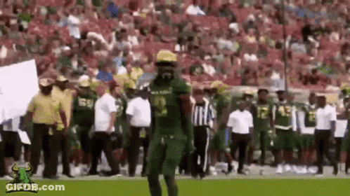 A Football Player In A Green Uniform Is Walking On A Field With A Referee . GIF