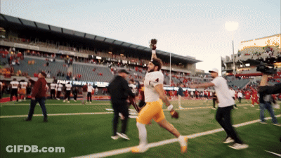 A Football Player Is Running On A Field With A Stadium In The Background That Says " Trophy Room " GIF