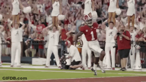 A Football Player Is Running On The Field With Cheerleaders Behind Him And A Crowd Watching . GIF