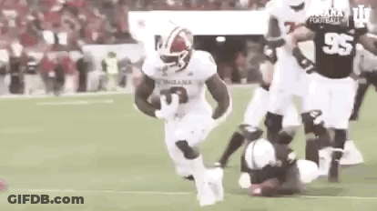 A Football Player Is Running With The Ball On A Field While A Player Is Laying On The Ground . GIF