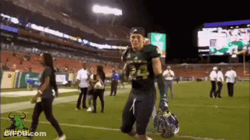 A Football Player Is Walking On A Field With A Helmet In His Hand . GIF