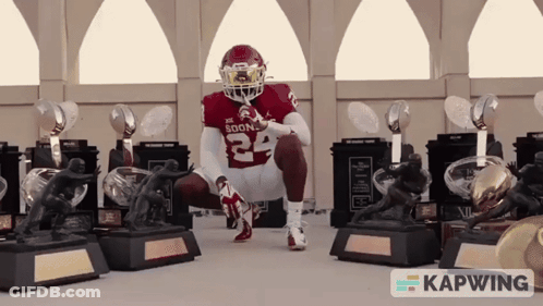A Football Player With The Number 24 On His Jersey Is Squatting Down In Front Of Trophies GIF