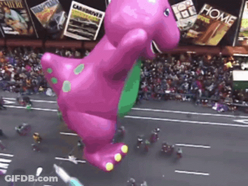 A Giant Balloon Of Barney The Dinosaur Is Flying Over A Street GIF