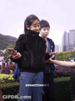 A Girl In A Black Sweater Is Holding A Cell Phone While Standing Next To A Boy In A Blue Jacket GIF