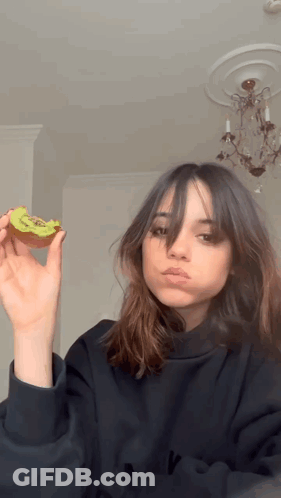 A Girl In A Black Sweatshirt Is Holding A Slice Of Kiwi In Her Hand GIF