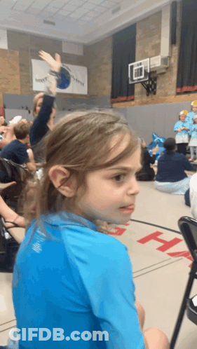 A Girl In A Blue Shirt Is Sitting In A Gym With A Basketball Hoop On The Wall GIF