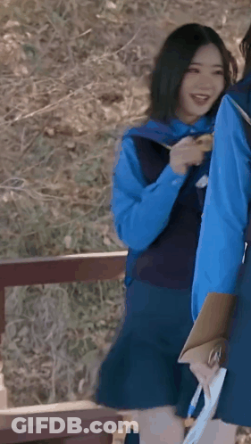 A Girl In A Blue Vest And Skirt Is Holding A Clipboard And Smiling While Standing Next To Another Girl . GIF