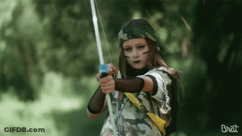 A Girl In A Camouflage Outfit Is Holding A Bow And Arrow And The Word Brat Is On The Bottom GIF