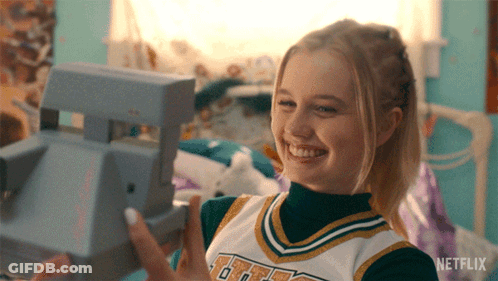 A Girl In A Cheerleader Uniform Is Holding A Polaroid Camera In Her Hand GIF