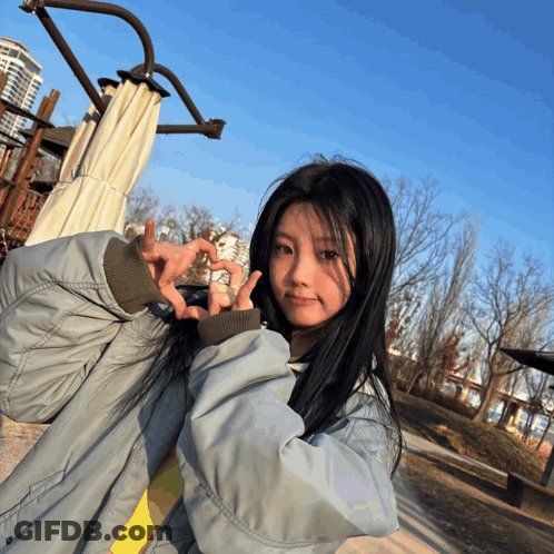 A Girl In A Grey Jacket Is Making A Heart With Her Hands GIF