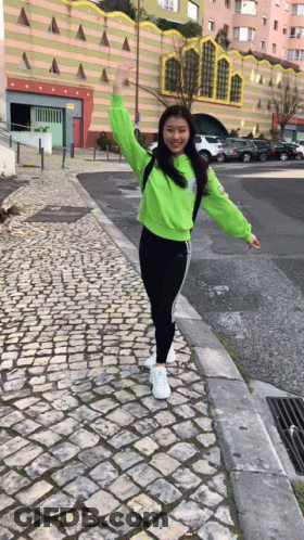 A Girl In A Neon Green Sweatshirt And Black Pants Is Standing On A Cobblestone Sidewalk . GIF