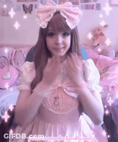 A Girl In A Pink Dress With A Pink Bow On Her Head Is Making A Heart With Her Hands . GIF