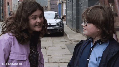A Girl In A Purple Jacket Stands Next To A Boy In A Blue Shirt With A License Plate That Says A72fyu GIF