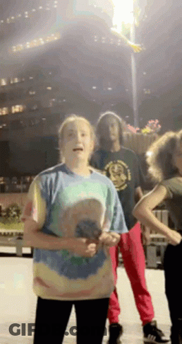 A Girl In A Tie Dye Shirt Stands In Front Of A Group Of People Dancing GIF