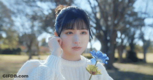 A Girl In A White Sweater Is Holding A Blue Flower In Her Hand . GIF