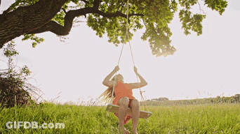 A Girl In An Orange Dress Is Sitting On A Wooden Swing Under A Tree GIF
